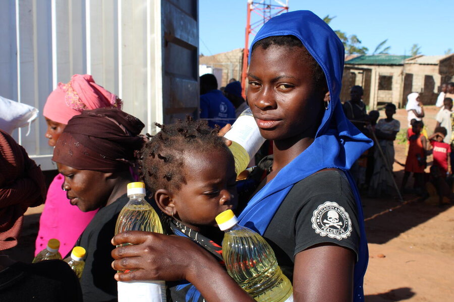 A woman with blue head and shoulders band carries two bottles of vegetable oil with her baby strapped to her fron 
